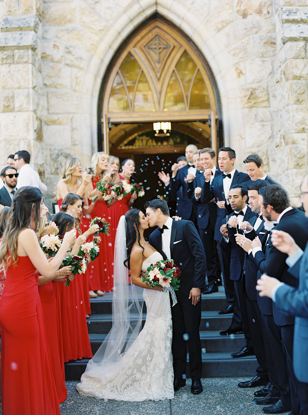surrounded by their friends, a bride and groom kiss in front of a church in Napa Valley, while bubbles float in the air. 